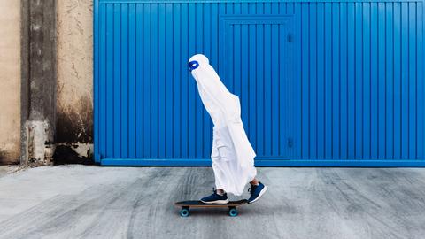 Ein Kind in einem Gespensterkostüm mit blauer Maske um die Augen fährt auf einem Skateboard an einem blauen Tor vorbei.