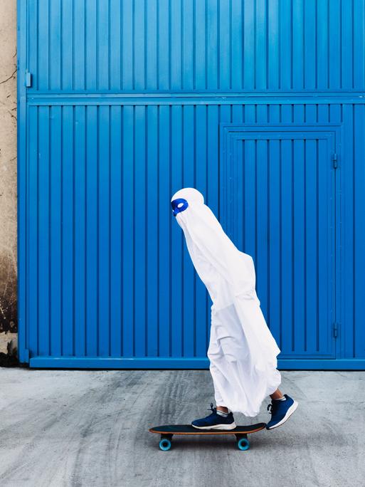 Ein Kind in einem Gespensterkostüm mit blauer Maske um die Augen fährt auf einem Skateboard an einem blauen Tor vorbei.
