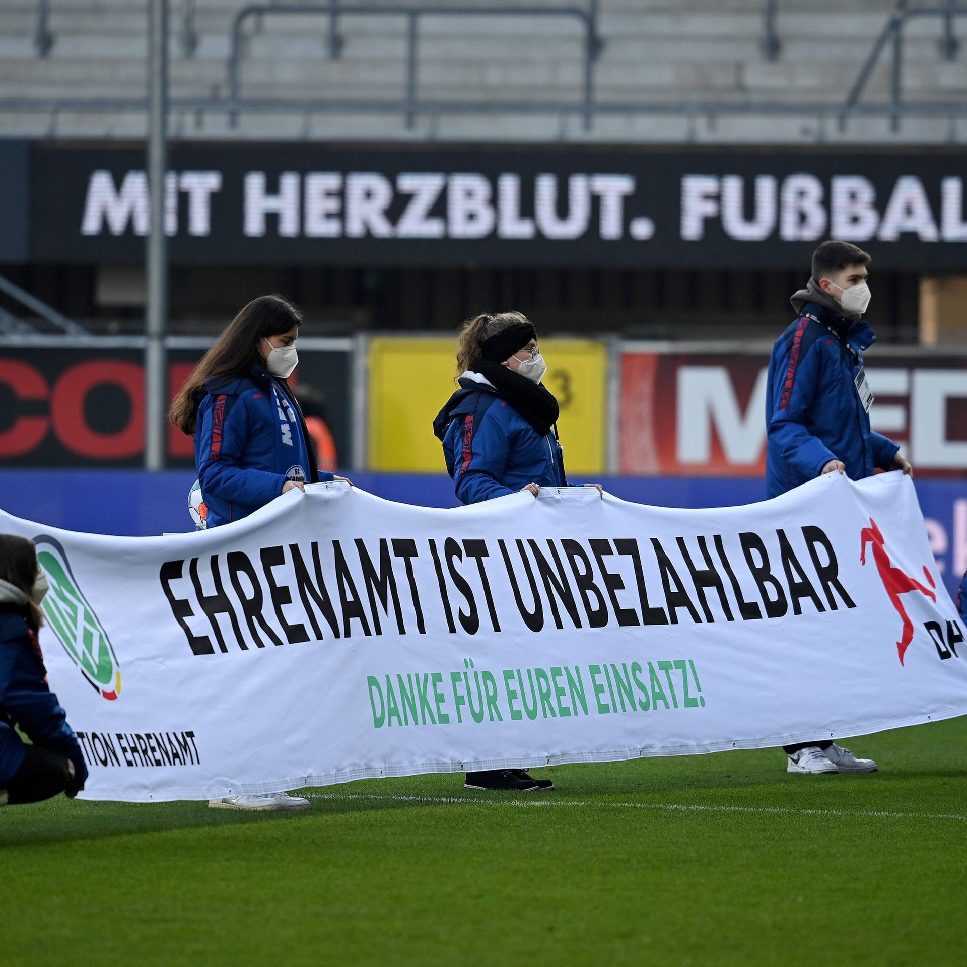 Freiwillige halten in einem Fußballstadion ein Banner hoch mit der Aufschrift: Ehrenamt ist unbezahlbar.
