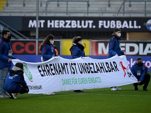 Freiwillige halten in einem Fußballstadion ein Banner hoch mit der Aufschrift: Ehrenamt ist unbezahlbar.
