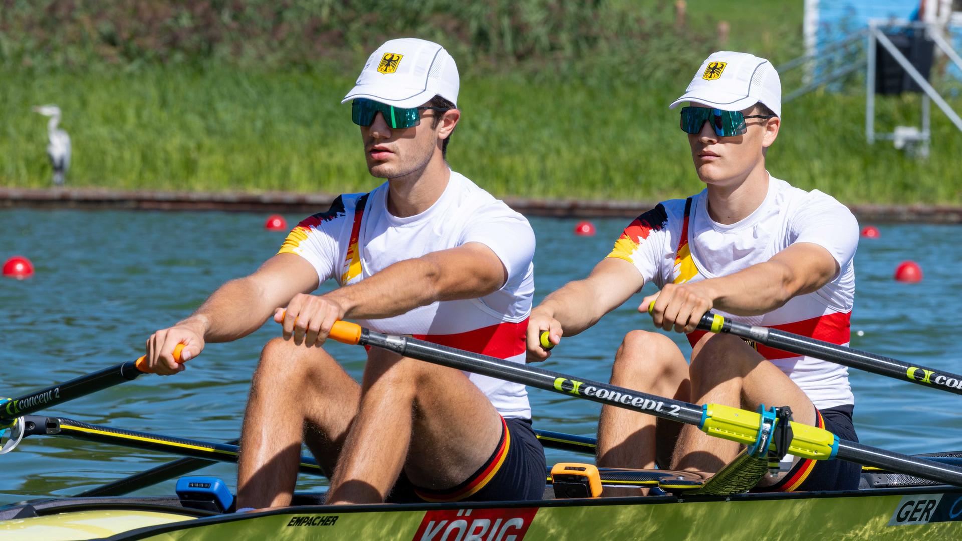 Die beiden in weißen Trikots sitzen auf dem Boot und rudern einen Kanal entlang. 