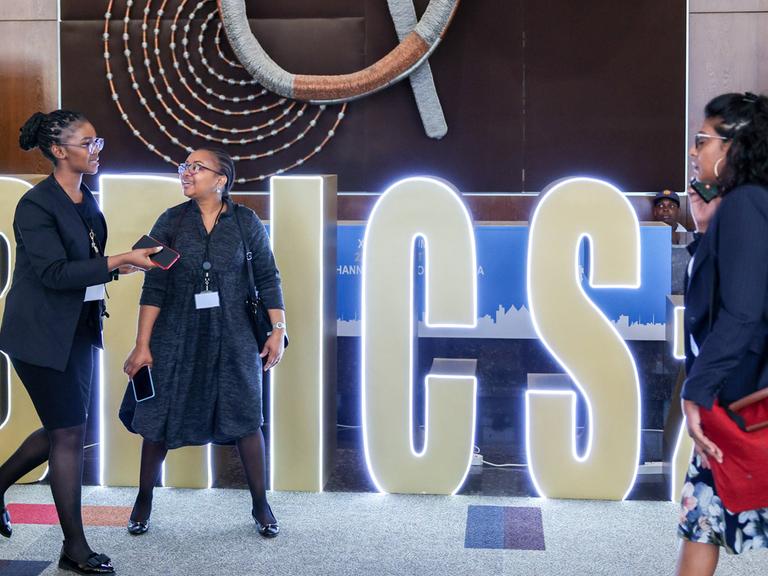 In einem Konferenzgebäude ist im Hintergrund der Schriftzug BRICS zu lesen; davor laufen drei Frauen vorbei und telefonieren teils; 22. August 2023