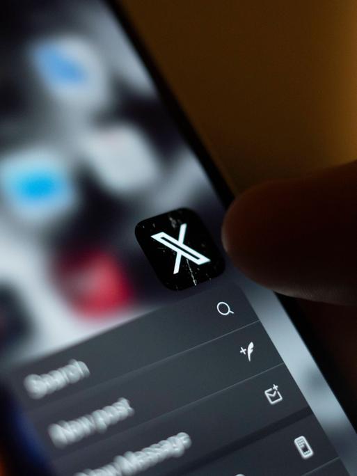 Auf einem Smartphone ist das Icon für die X-App (ehemals Twitter) zu sehen. Eine Hand hält das Handy und der Daumen zielt auf die App.