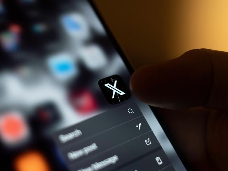 Auf einem Smartphone ist das Icon für die X-App (ehemals Twitter) zu sehen. Eine Hand hält das Handy und der Daumen zielt auf die App.