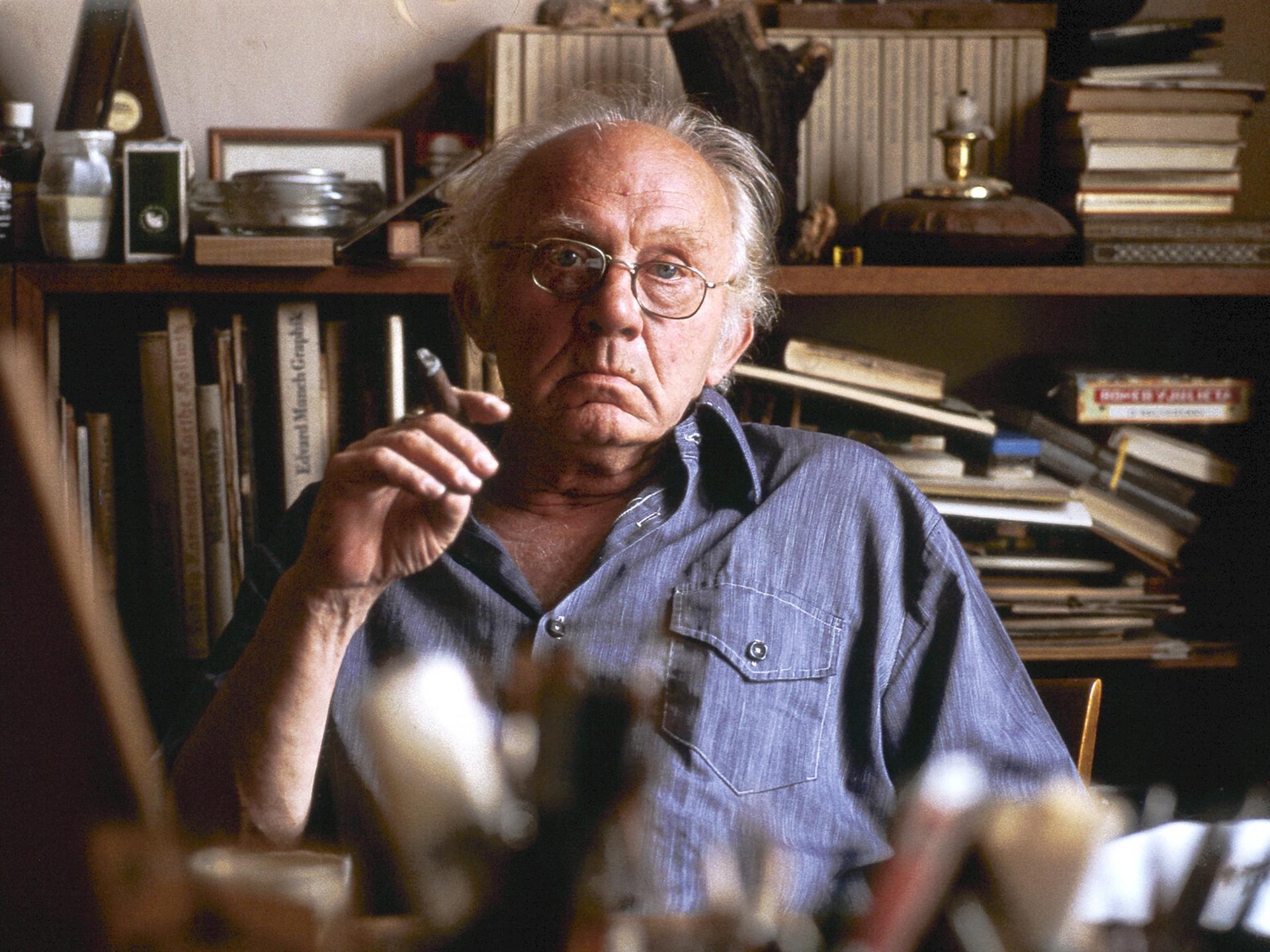 Der Berliner Maler und Grafiker Arno Mohr sitzt im Jahr 1978 mit einer Zigarre in der Hand vor einer Regalwand in seiner Wohnung in Berlin.