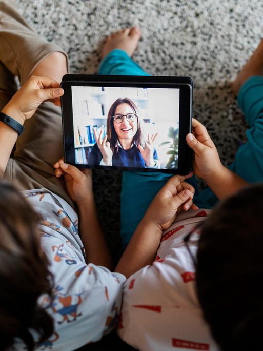 Zwei Brüder sitzen zu Hause auf dem Boden und nutzen ein digitales Tablet für einen Videochat mit ihrer Mutter. 