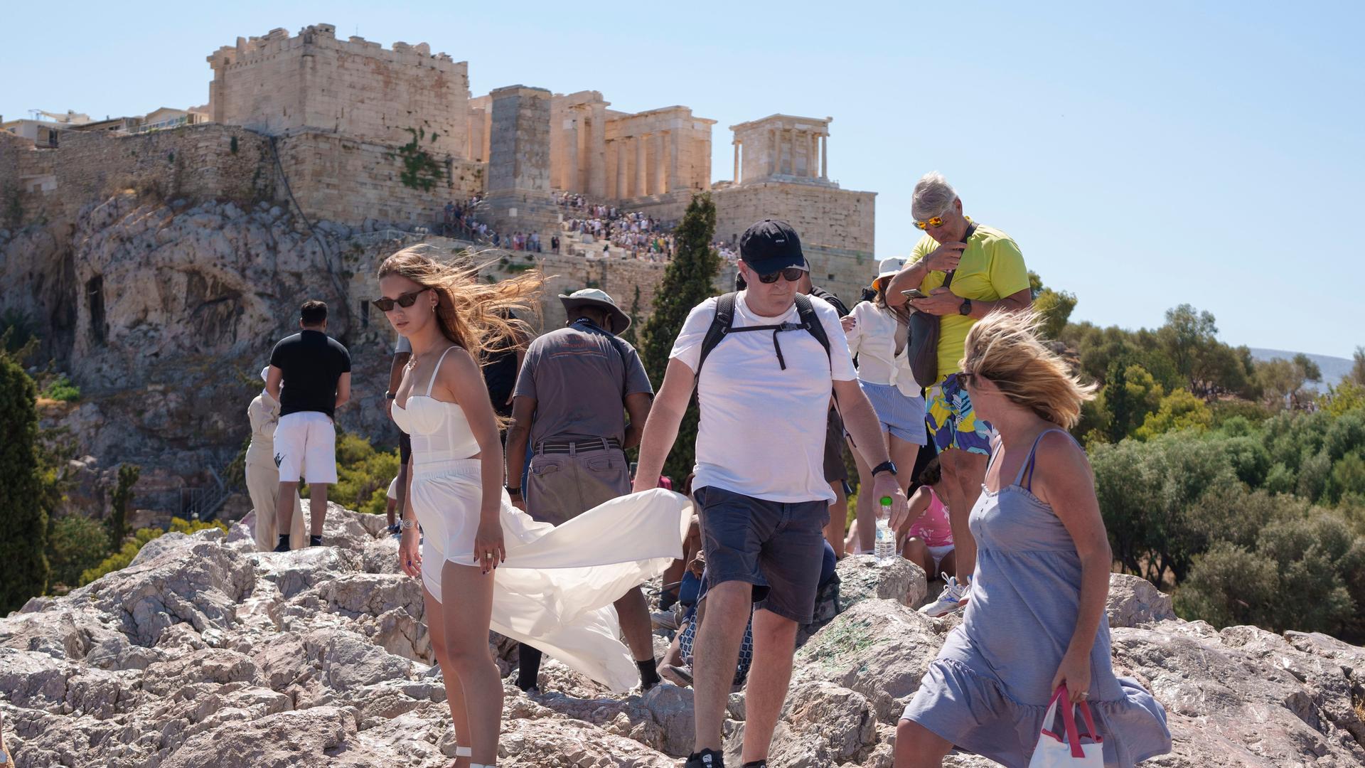 Touristen gehen an einem heißen Tag auf dem Areopagous-Hügel, während im Hintergrund der antike Akropolis-Hügel steht. 
