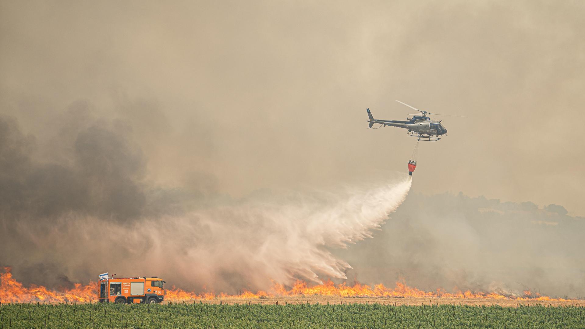 Ein israelischer Löschhubschrauber schüttet Wasser auf ein Feuer am Rande einer Plantage. Links im Bild steht ein großes Löschfahrzeug der Feuerwehr. Im Hintergrund sind Flammen und starker Rauch zu sehen. Der Himmel ist kaum zu erkennen. 