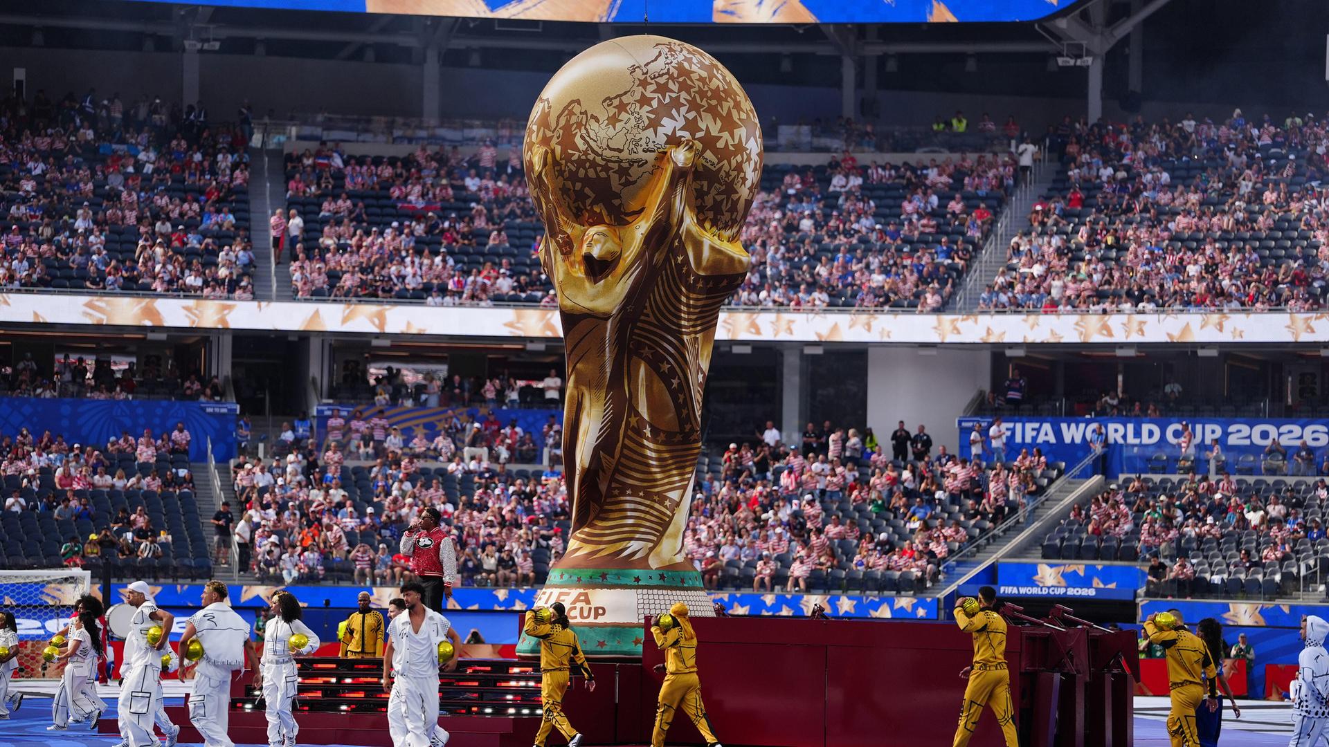 Die Fußball-Weltmeisterschaften 2026 finden in mehreren Ländern statt. Ein riesiger Pokal ist auf dem Spielfeld, viele Menschen sitzen im Publikum und feiern auf dem Platz. 