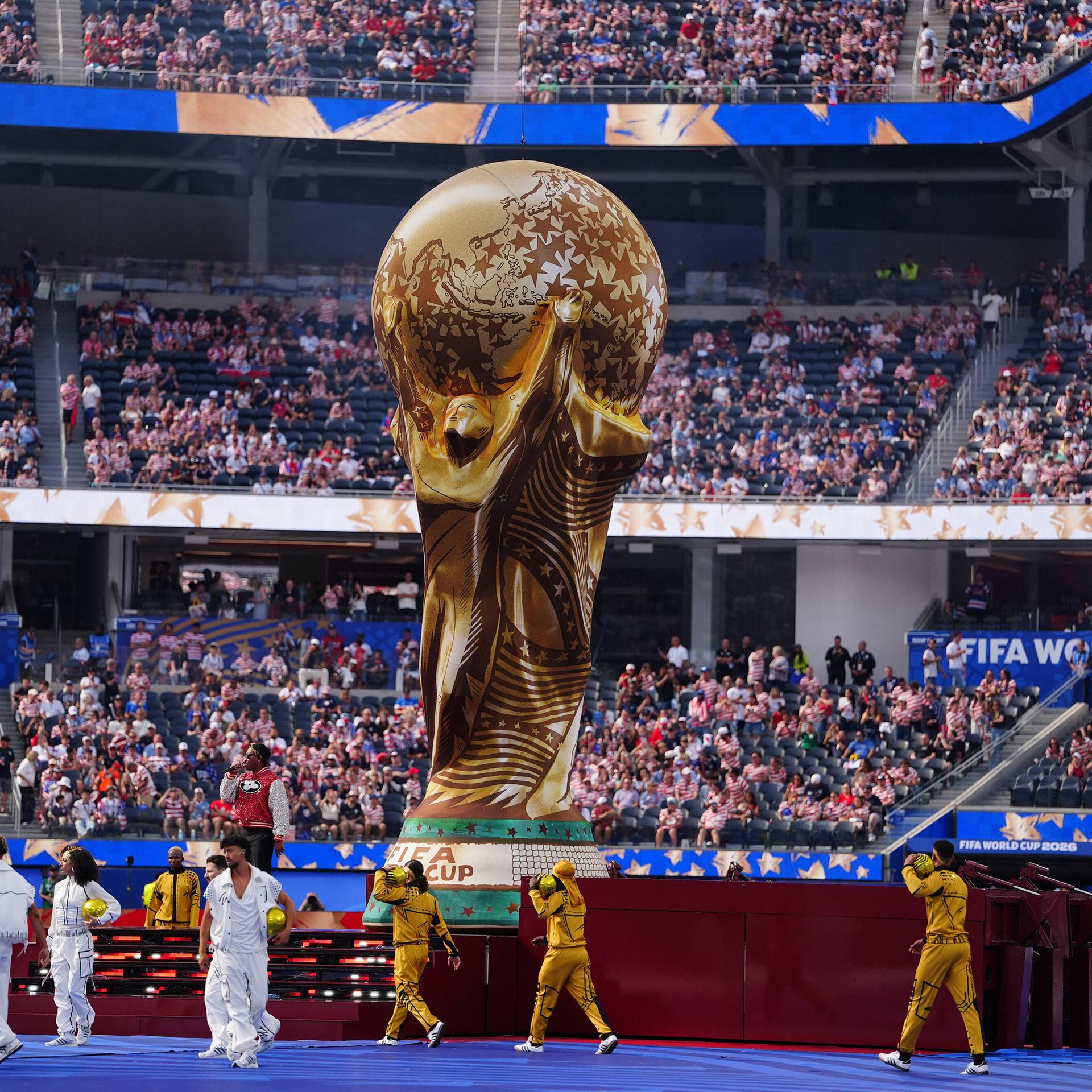 Die Fußball-Weltmeisterschaften 2026 finden in mehreren Ländern statt. Ein riesiger Pokal ist auf dem Spielfeld, viele Menschen sitzen im Publikum und feiern auf dem Platz. 