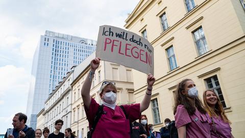 Eine Frau in berufsspezifischer Kleidung hält ein Transparent hoch, auf dem "Ich will doch nur pflegen" steht. 