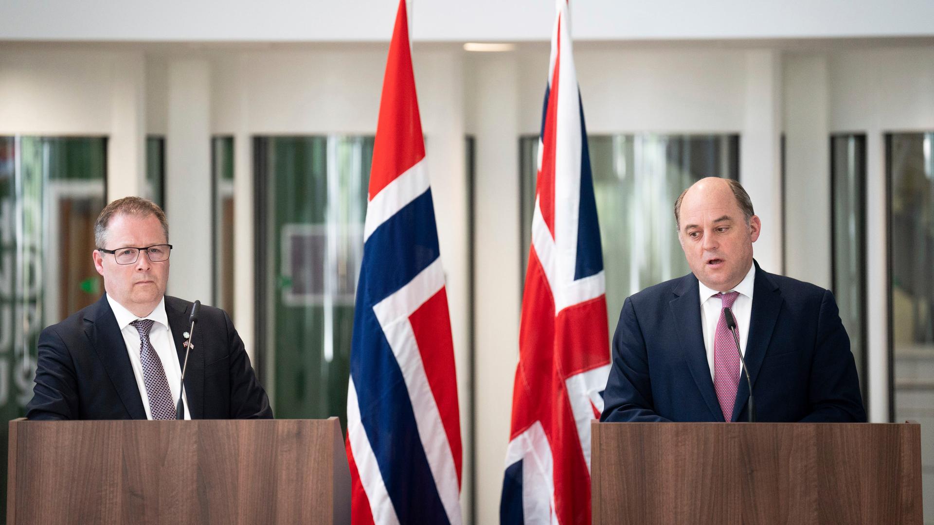 Die beiden stehen nebeneinander an hölzernen Rednerpulten; Wallace sagt etwas. Zwischen ihnen zwei britische Flaggen. Die beiden stehen nebeneinander an hölzernen Rednerpulten; Wallace sagt etwas. Zwischen ihnen zwei britische Flaggen.