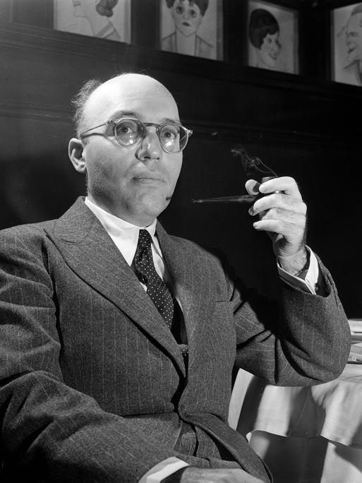 Kurt Weill sitzt mit einer Pfeife an einem Tisch in einem Salon.