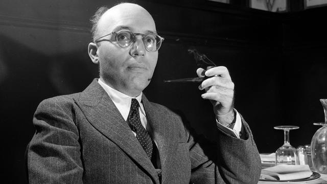 Kurt Weill sitzt mit einer Pfeife an einem Tisch in einem Salon.