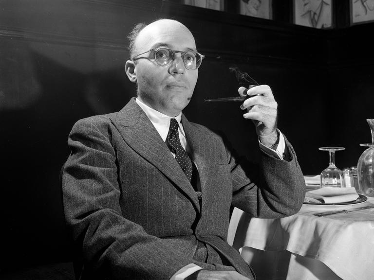 Kurt Weill sitzt mit einer Pfeife an einem Tisch in einem Salon.