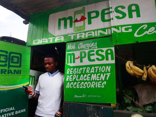 Kleine Hütte mit der Aufschrift M-Pesa, in deren Tür ein Mann mit Smartphone in der Hand steht.
