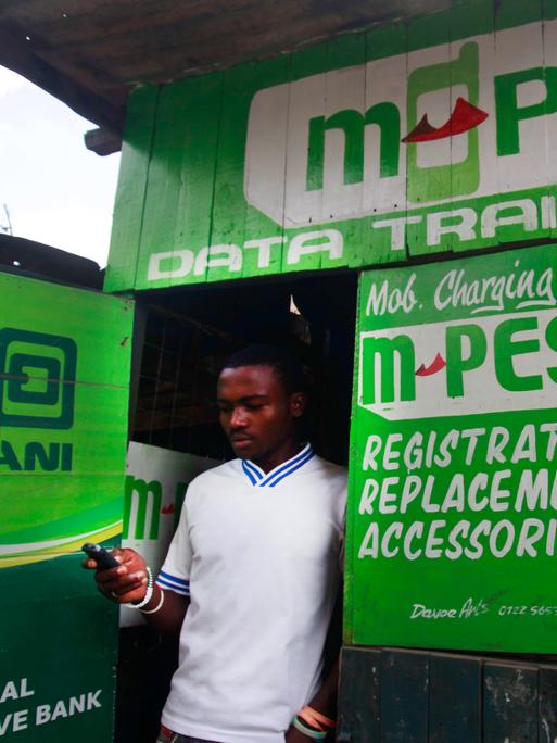 Kleine Hütte mit der Aufschrift M-Pesa, in deren Tür ein Mann mit Smartphone in der Hand steht.