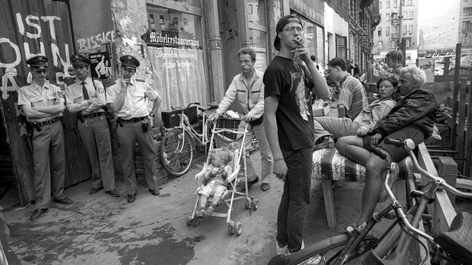 Eine Straßenszene mit Berlin mit einem rauchenden Mann im Vordergrund, Polizisten, die an einem Eingang lehnen und einem Mann, der einen Kinderwagen an Straßencafes vorbeischiebt. 