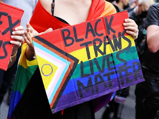 Demoteilnehmerin mit umgehangener Regenbogenfahne trägt Protestschild mit der Aufschrift: Black Trans Lives Matter