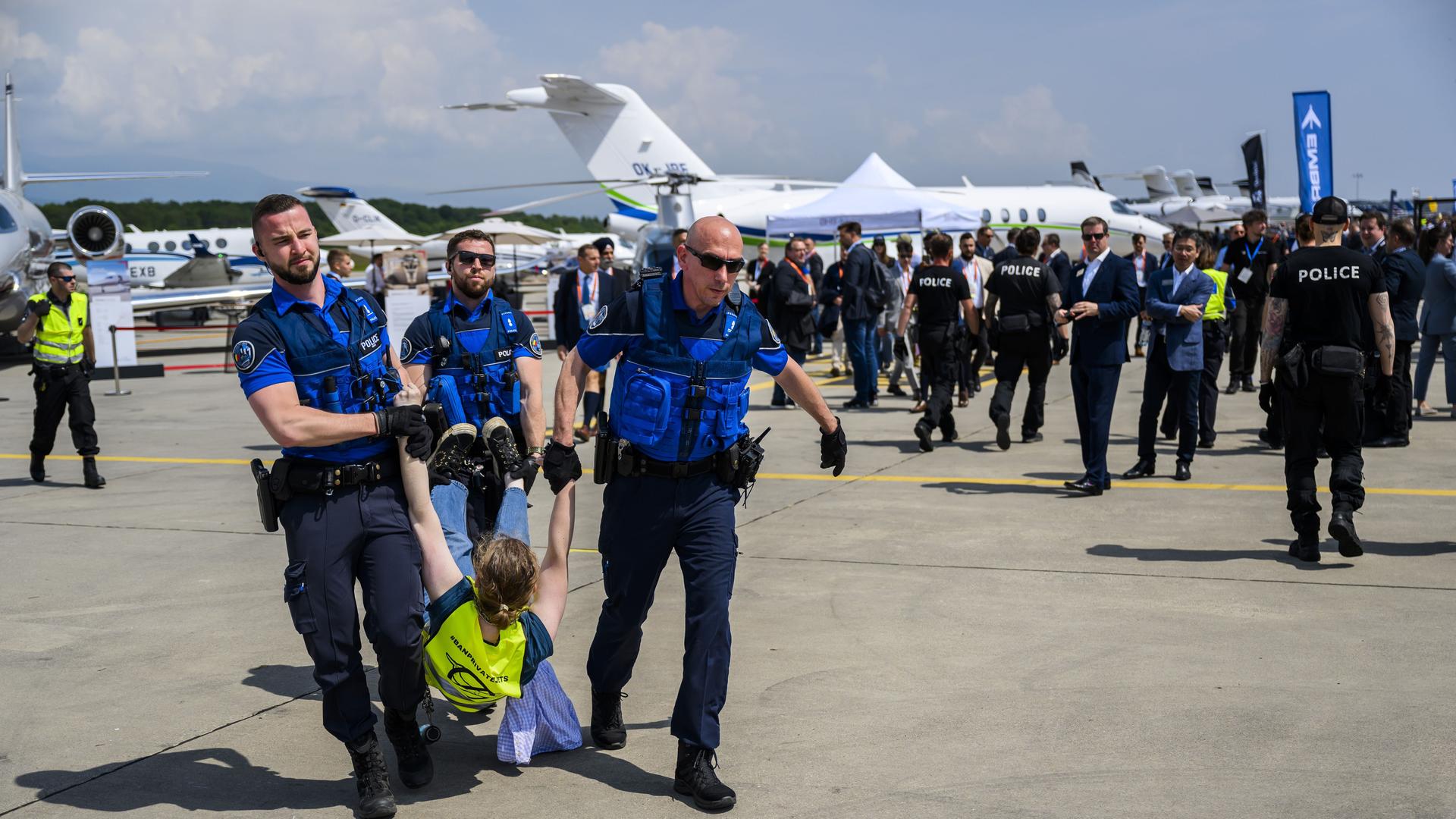 Drei Polizisten tragen einen Klimaaktivsten in Genf am Flughafen vom Rollfeld. Im Hinergrund stehen Flugzeuge und viele Polizisten.