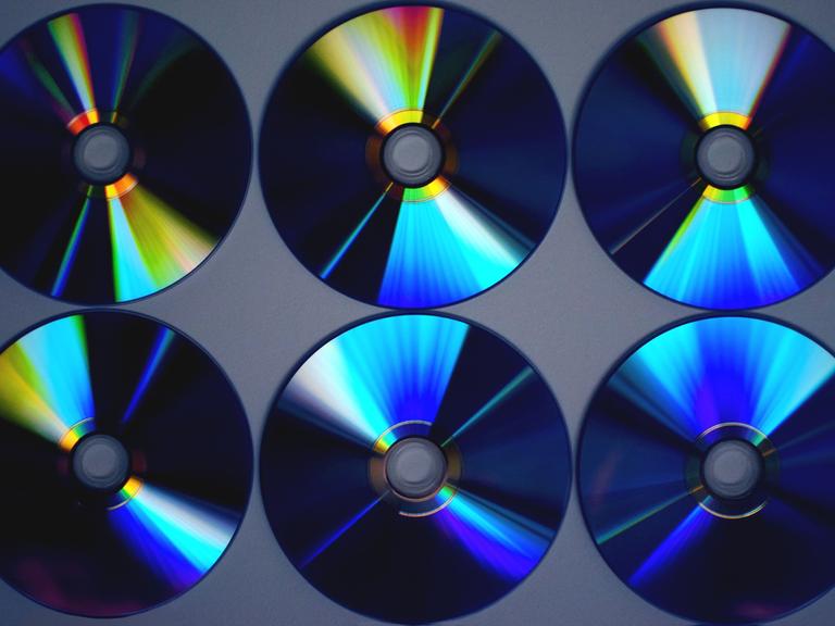 Auf dem Bild sind sechs CDs zu sehen, nicht mit der Cover-Seite, sondern die mit Inhalt bespielte Seite. Die CD-Ringe glänzen in blauen und gelben Farben.