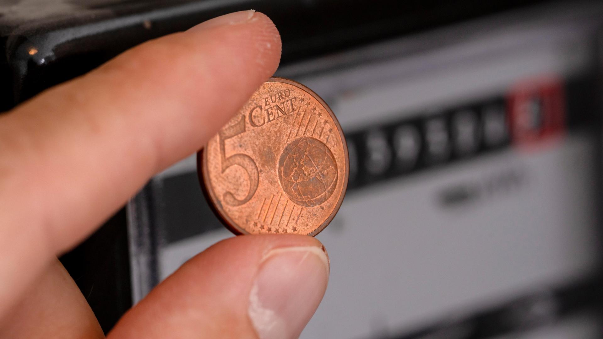 Hand hält eine 5 Euro Cent Münze vor Stromzähler. 