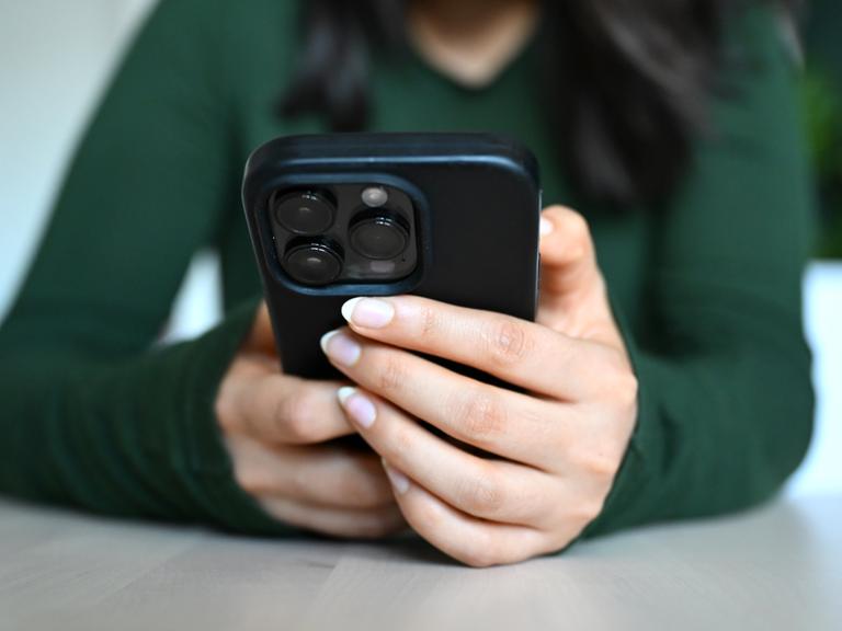 Hände einer jungen Frau umgreifen ein Smartphone an einem Tisch.