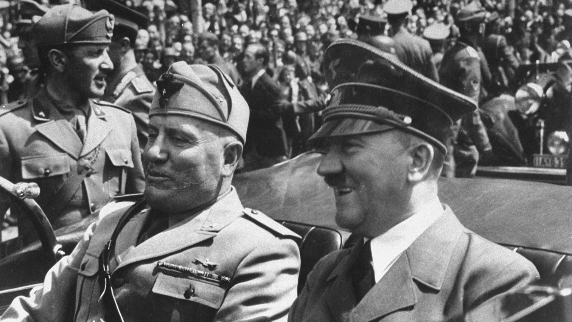 Schwarz-weiss Bild von Adolf  Hitler  (R)  und  Benito  Mussolini  bei einer Fahrt im Cabrio. Im Hintergrund viele Menschen.