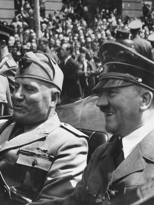 Schwarz-weiss Bild von Adolf  Hitler  (R)  und  Benito  Mussolini  bei einer Fahrt im Cabrio. Im Hintergrund viele Menschen.