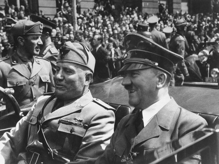 Schwarz-weiss Bild von Adolf  Hitler  (R)  und  Benito  Mussolini  bei einer Fahrt im Cabrio. Im Hintergrund viele Menschen.