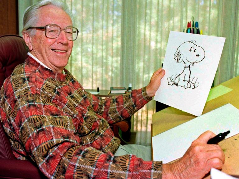 Charles Schulz hält eine Zeichnung von Snoopy in der Hand und lächelt.