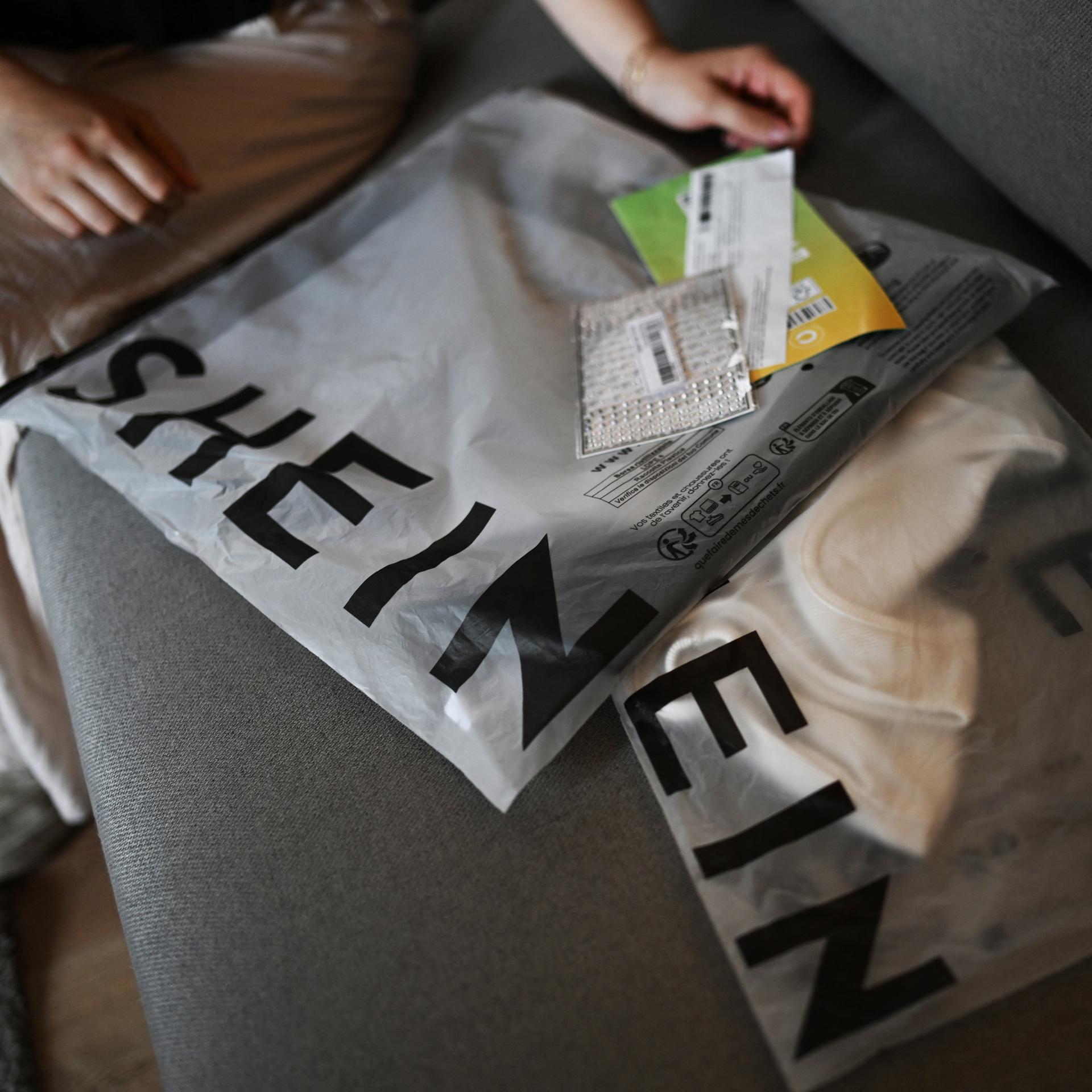 Päckchen mit der Aufschrift "Shein" mit darin enthaltener Kleidung sind auf einem Sofa ausgebreitet.