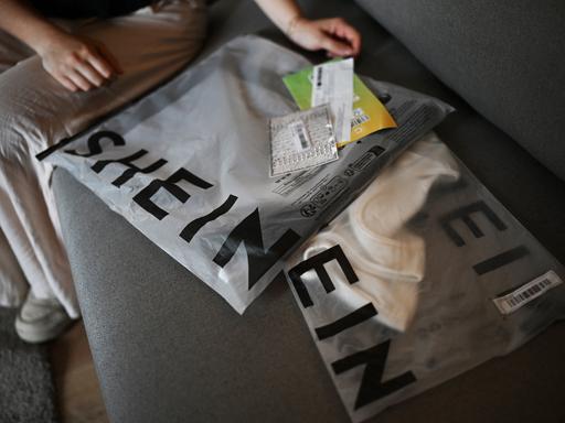 Päckchen mit der Aufschrift "Shein" mit darin enthaltener Kleidung sind auf einem Sofa ausgebreitet.