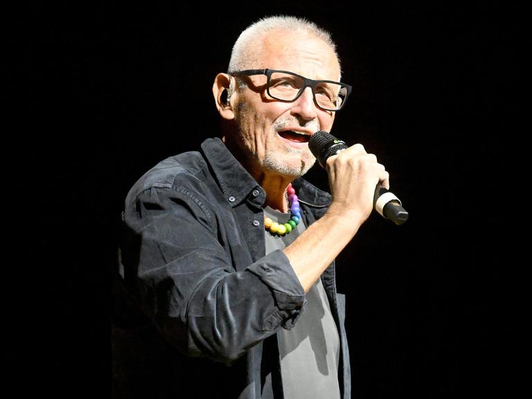 Der Liedermacher Konstantin Wecker steht auf einer Bühne und singt in ein Mikrofon.