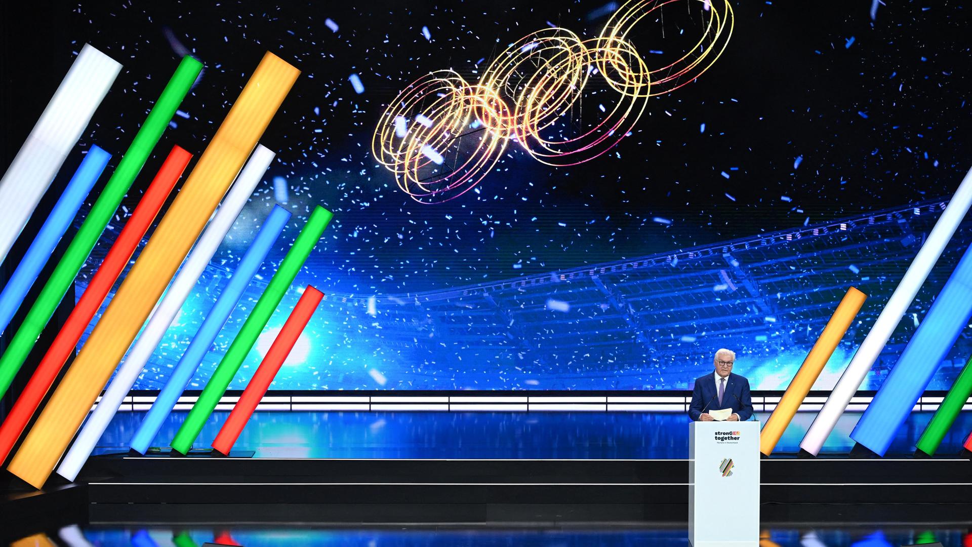 Bundespräsident Steinmeier spricht auf einer Bühne. Im Hintergrund erscheinen die olympischen Ringe auf einer Leinwand.
      