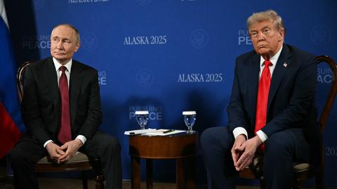 Putin und Trump sitzen auf Stühlen und schauen ernst an der Kamera vorbei. Zwischen ihnen steht ein kleiner Tisch. 