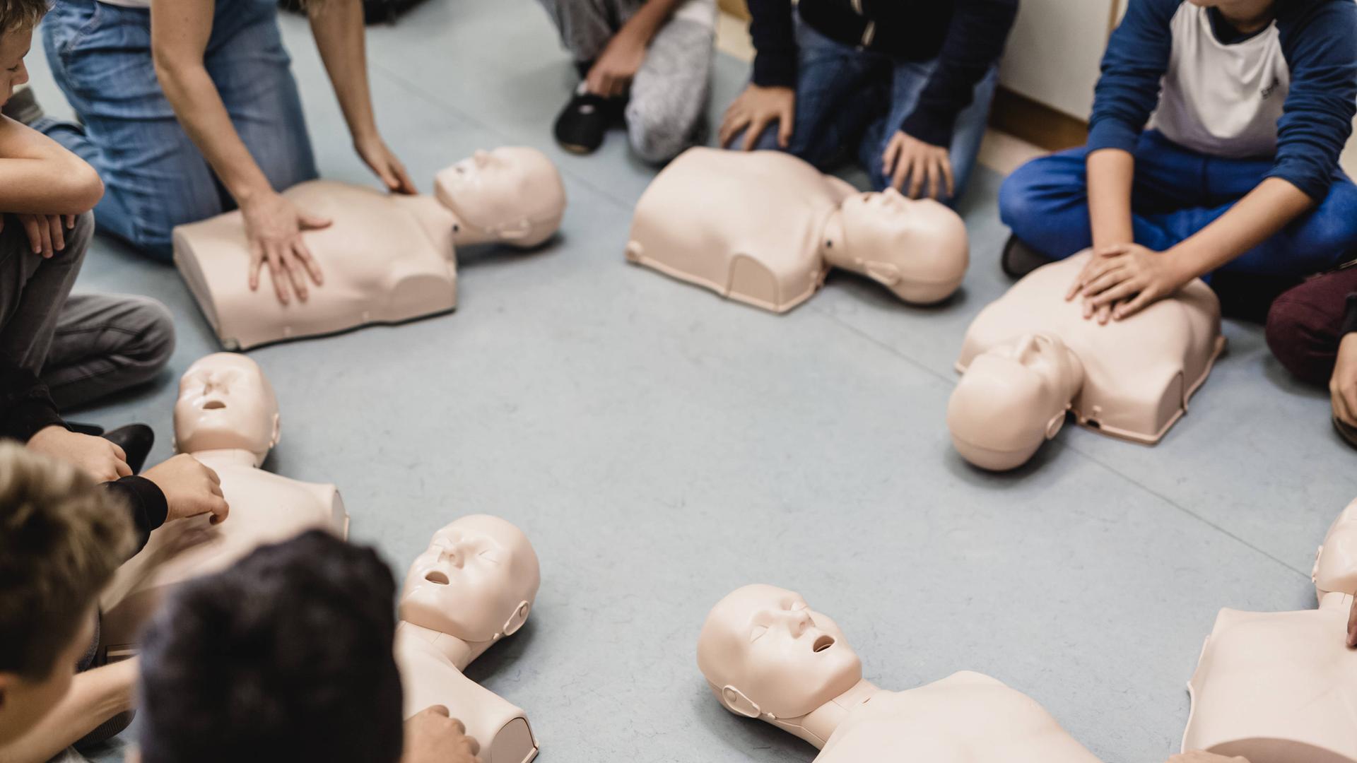 Mehrere Menschen sitzen im Kreis und üben die Reanimation an Plasikpuppen, die vor ihnen auf dem Boden liegen. 