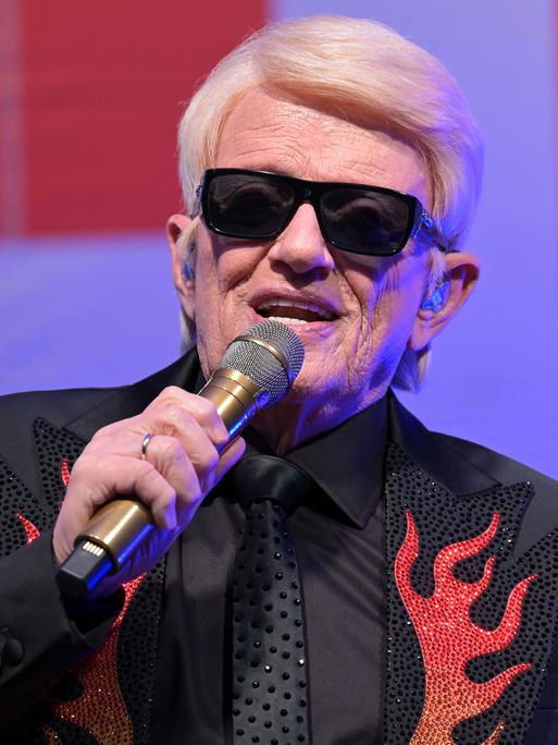 Nahaufnahme vom Oberkörper des blondhaarigen Sängers mit der markanten schwarzen Sonnenbrille, der gerade in ein Mikrofo singt. Er träg ein schwarzes Jackett mit roten flammenartigen Applikationen. 