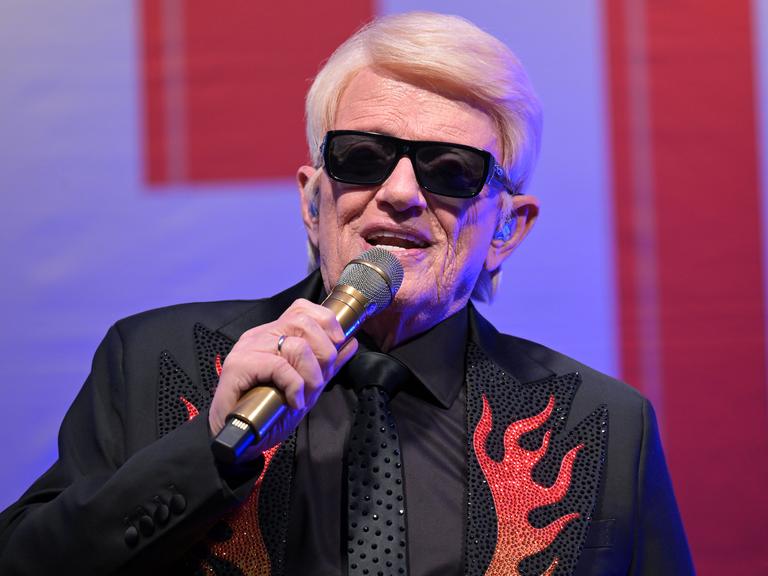 Nahaufnahme vom Oberkörper des blondhaarigen Sängers mit der markanten schwarzen Sonnenbrille, der gerade in ein Mikrofo singt. Er träg ein schwarzes Jackett mit roten flammenartigen Applikationen. 