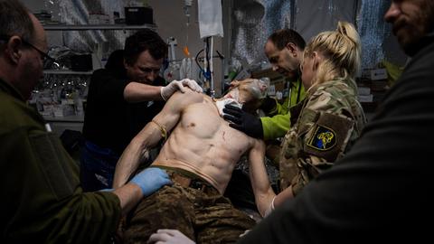 Ein verwundeter Mann ist umgeben von Ärzten und Pflegern, die ihn versorgen. Er trägt eine Camouflage-Hose, ebenso wie eine Frau, die rechts von ihm steht mit dem Rücken zur Kamera. Der Oberkörper des Mannes ist frei.
