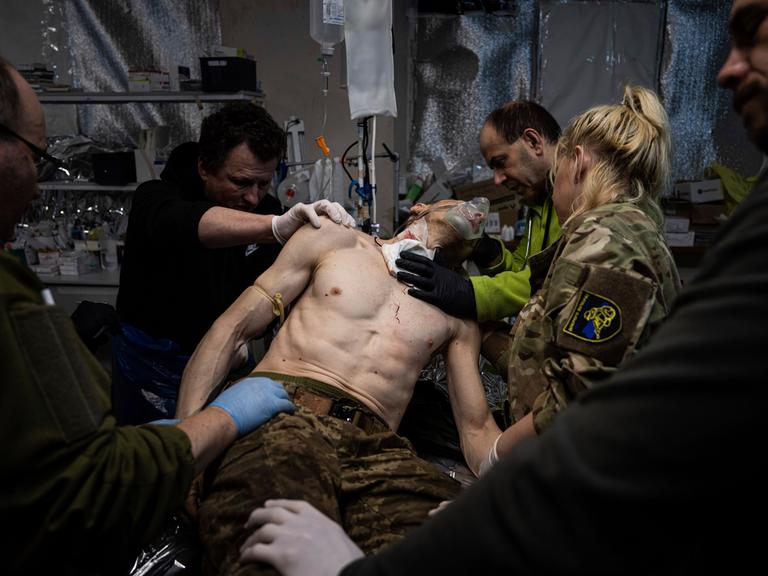 Ein verwundeter Mann ist umgeben von Ärzten und Pflegern, die ihn versorgen. Er trägt eine Camouflage-Hose, ebenso wie eine Frau, die rechts von ihm steht mit dem Rücken zur Kamera. Der Oberkörper des Mannes ist frei.