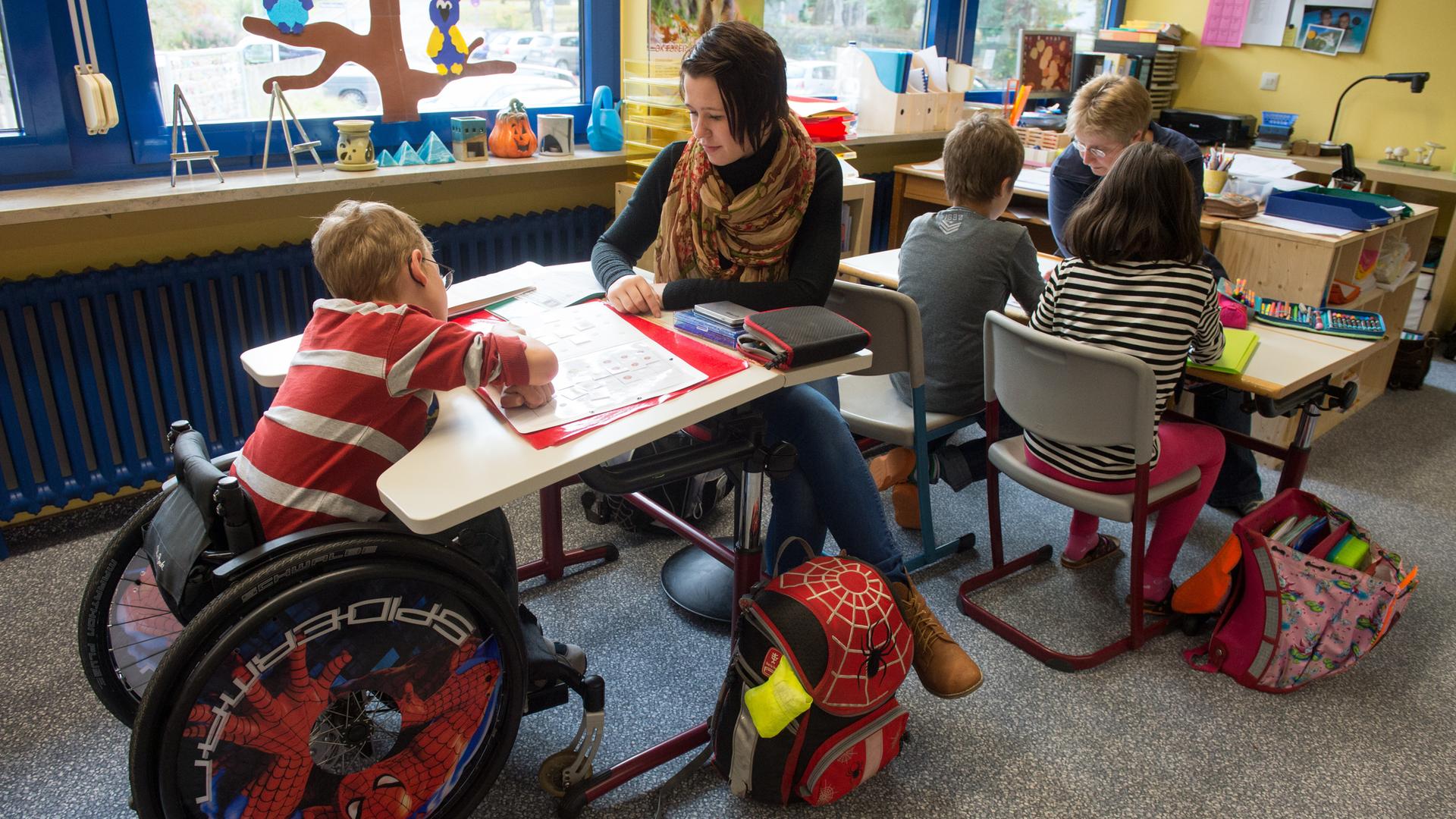 Zwei Frauen sitzen mit Schul-Kindern an Tischen in einem Klassen-Raum. Ein Kind sitzt im Roll-Stuhl. Zwei Frauen sitzen mit Schul-Kindern an Tischen in einem Klassen-Raum. Ein Kind sitzt im Roll-Stuhl.