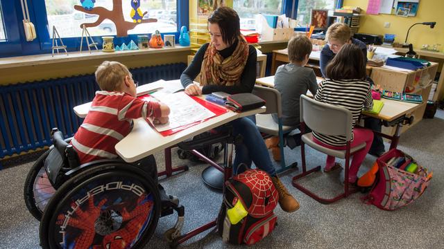 Zwei Frauen sitzen mit Schul-Kindern an Tischen in einem Klassen-Raum. Ein Kind sitzt im Roll-Stuhl.