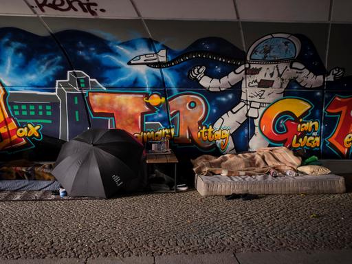 Unter einer mit Graffiti bemalten Brücke befindet sich das Lager eines Obdachlosen mit Matratzen, aufgespanntem Regenschirm und einem faltbaren Tisch.