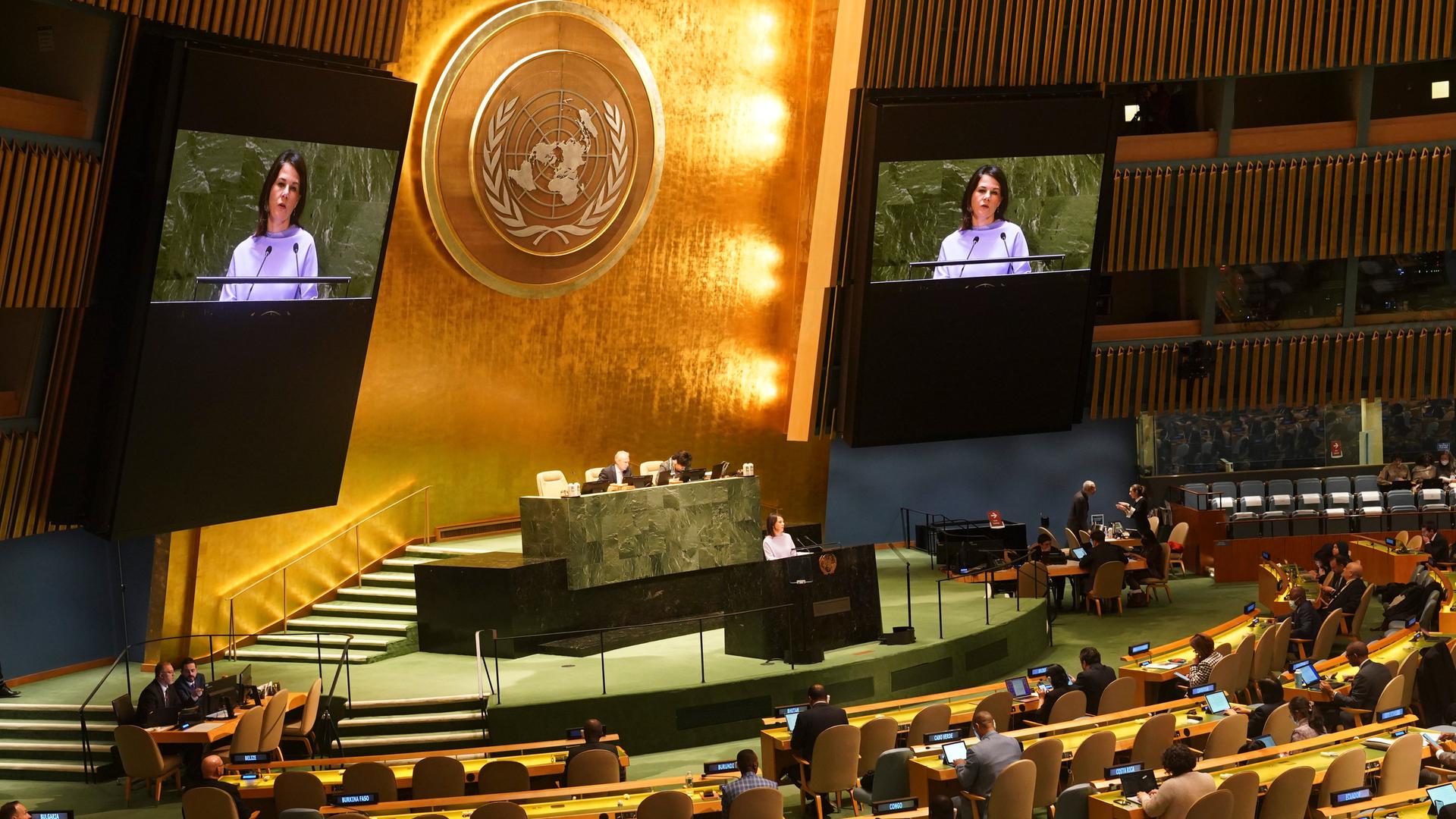 Außenministerin Baerbock vor der Vollversammlung der Vereinten Nationen: sie ist am Pult zu sehen, über ihr zwei große Monitore, die ihr Gesicht zeigen, in der Mitte das Logo der Vereinten Nationen in Gold.