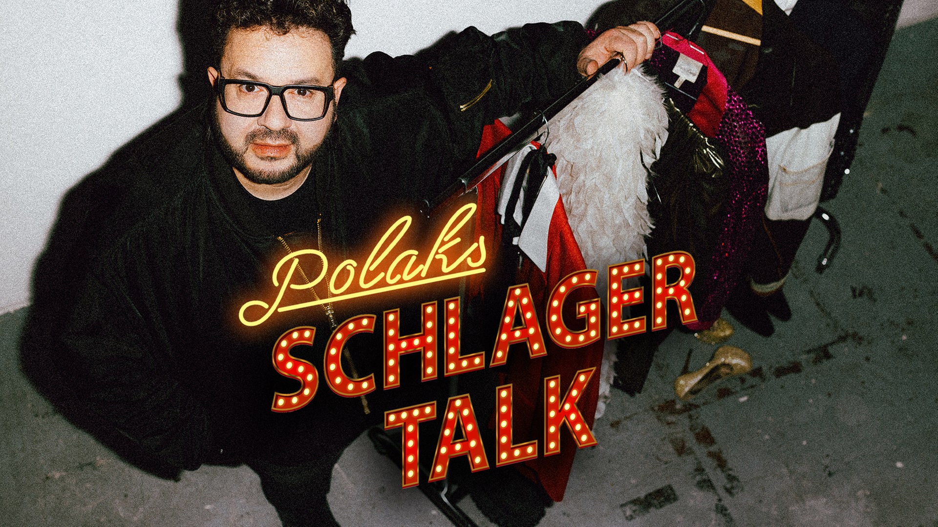 Polaks Schlagertalk: Podcast-Host Oliver Polak steht neben einer Garderobenstange mit bunten Bühnenoutfits wie Federboa und Glitzeranzügen