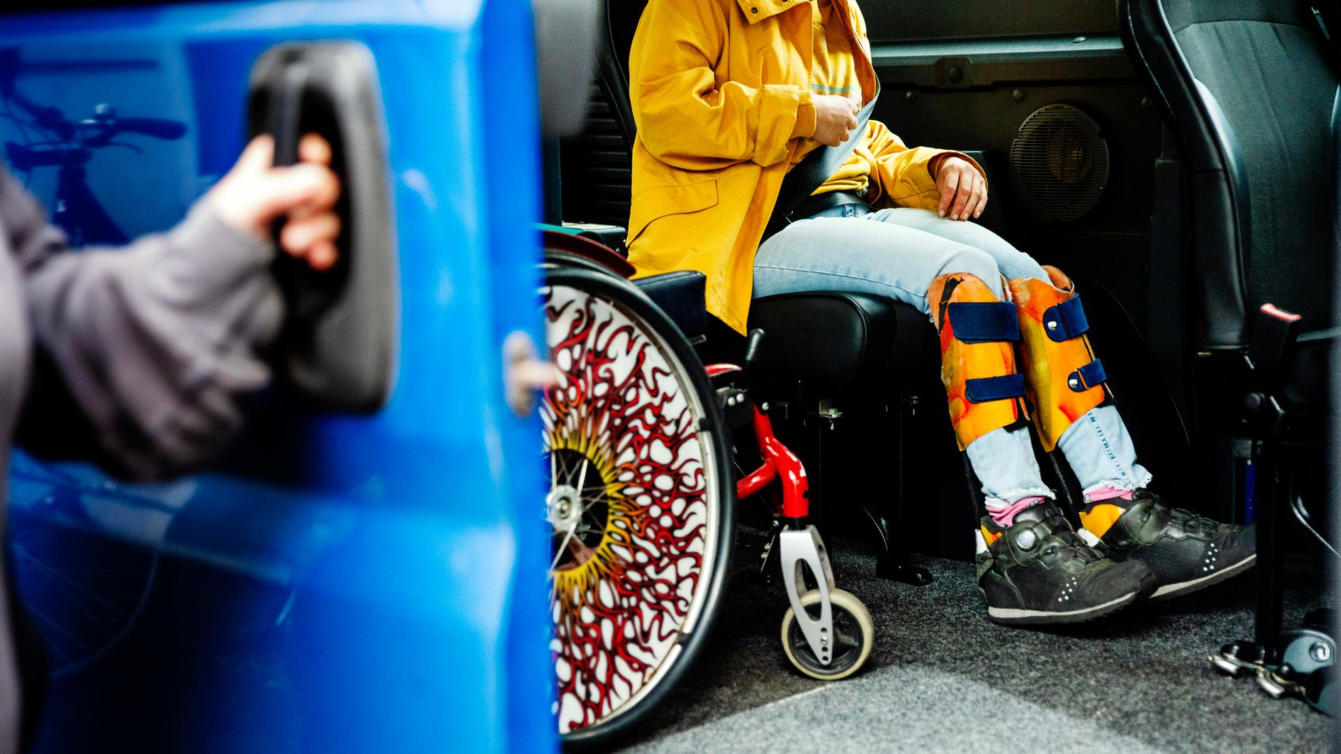 Eine junge behinderte Frau sitzt angeschnallt im Fahrdienstwagen, neben ihr parkt der Rollstuhl. Die Tür wird vom Fahrdienst zugezogen. Berlin, 2024