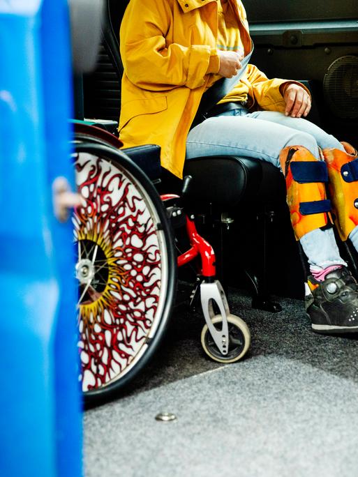 Eine junge behinderte Frau sitzt angeschnallt im Fahrdienstwagen, neben ihr parkt der Rollstuhl. Die Tür wird vom Fahrdienst zugezogen. Berlin, 2024