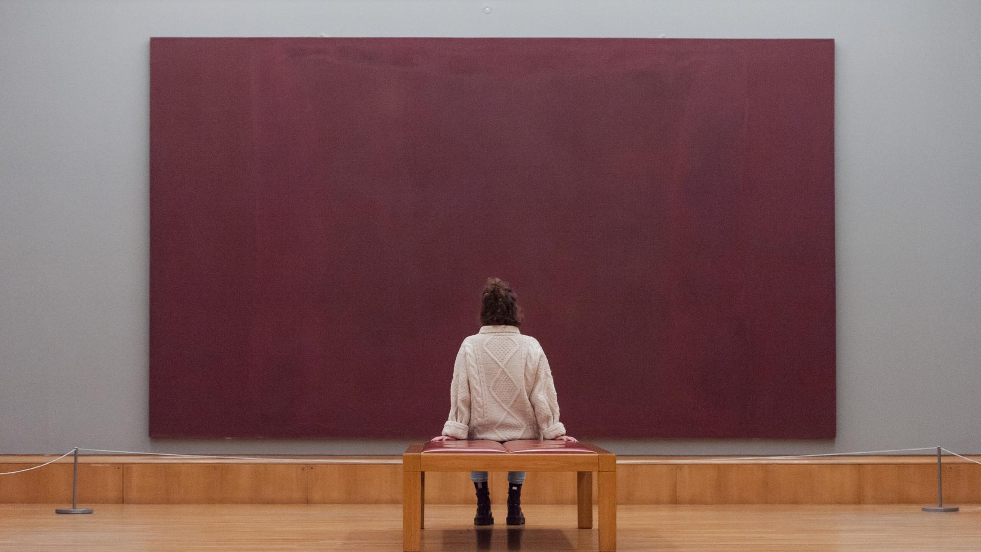 Eine Frau blickt auf einer Bank sitzend auf das Bild "Red on Maroon" (1959) aus der Reihe The Seagram Murals von Mark Rothko Eine Frau blickt auf einer Bank sitzend auf das Bild "Red on Maroon" (1959) aus der Reihe The Seagram Murals von Mark Rothko