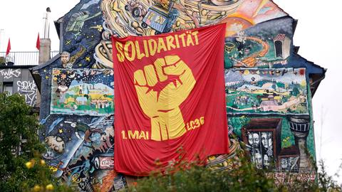 An einer hohen Hauswand, die mit zahlreichen Szenen bemalt wurde, hängt ein rotes Tuch. Darauf sieht man eine geballte Faust und die Worte "Solidarität 1. Mai".